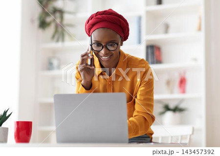Smiling Black Woman Using Laptop And Holding Phone While Engaging In Work, Sitting At Home Office Desk. Focused And Productive Environment, Business Concept. 143457392