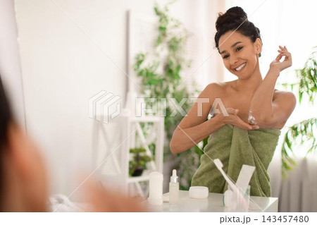 Smiling Young Woman Applying Moisturizer On Her Arm While Looking In Mirror, Sitting At Home. She Enjoys A Skincare Routine For Healthy Skin In A Bright, Relaxing Bathroom Setting. Smiling Young Woman Applying Moisturizer On Her Arm While Looking In Mirror, Sitting At Home. She Enjoys A Skincare Routine For Healthy Skin In A Bright, Relaxing Bathroom Setting. 143457480