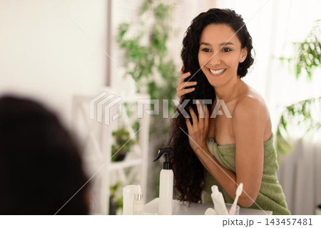 Smiling Young Woman With Curly Hair Applying Hair Product In Front Of Mirror At Home, Taking Care Of Her Hair And Wellness. Home Setting, Beauty And Self-care Concept. Smiling Young Woman With Curly Hair Applying Hair Product In Front Of Mirror At Home, Taking Care Of Her Hair And Wellness. Home Setting, Beauty And Self-care Concept. 143457481