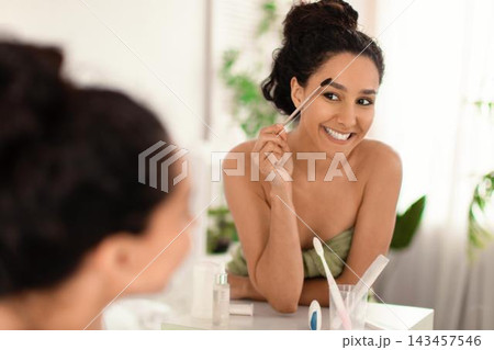 Smiling Young Woman With Natural Beauty Holding Eyebrow Brush While Looking At Mirror In Cozy Bathroom. Engaged In Self-care Routine, Enjoying Skincare At Home, Beauty Concept. 143457546