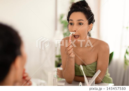 Focused Young Woman Of Middle Eastern Descent Engaged In Hair Removal By Shaving Upper Lip With Epilator In Front Of Mirror At Home, Emphasizing Personal Grooming Routine. Beauty Concept. 143457580