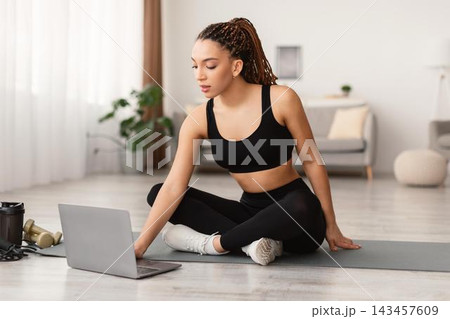 Focused Young Black Woman Sitting Cross-legged On Exercise Mat, Engaging In Online Fitness Routine At Home, Using Laptop, Promoting Healthy Lifestyle And Wellbeing. Fitness Concept. Focused Young Black Woman Sitting Cross-legged On Exercise Mat, Engaging In Online Fitness Routine At Home, Using Laptop, Promoting Healthy Lifestyle And Wellbeing. Fitness Concept. 143457609