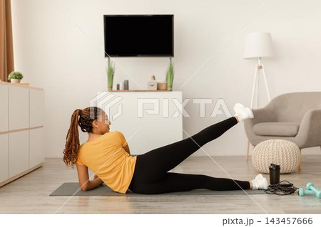 Focused Black Female Exercising At Home On Yoga Mat, Doing Side Leg Raises In Modern Living Room. Staying Active And Fit With Home Workouts, Emphasizing Importance Of Fitness Concept. Focused Black Female Exercising At Home On Yoga Mat, Doing Side Leg Raises In Modern Living Room. Staying Active And Fit With Home Workouts, Emphasizing Importance Of Fitness Concept. 143457666
