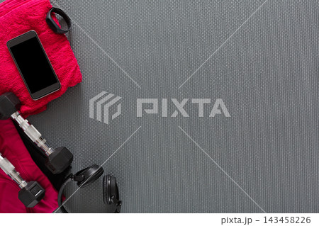 A setup shows a smartphone, headphones, dumbbells, and a pink towel on a gray gym mat. This arrangement highlights typical items needed for an exercise routine. 143458226