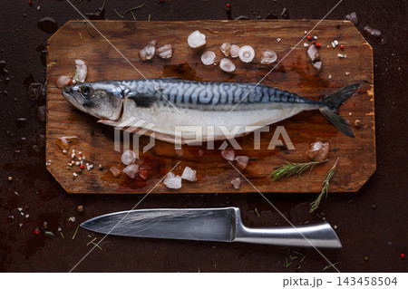A whole mackerel fish sits on a wooden cutting board with ice and spices around it. A sharp knife is placed next to the fish. This setup is typical for meal preparation. 143458504