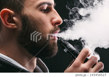 A person with a beard uses a vape device, producing a thick cloud of vapor. The setting is dark, emphasizing the vapor's visibility as it disperses into the air. A person with a beard uses a vape device, producing a thick cloud of vapor. The setting is dark, emphasizing the vapor's visibility as it disperses into the air. 143458527