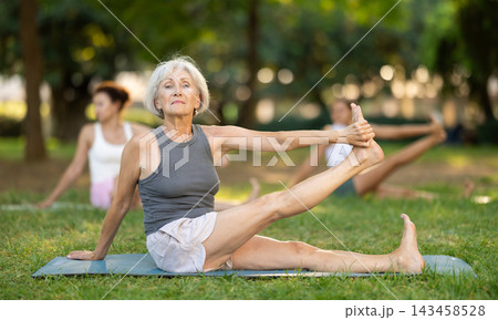 Concentrated mature lady making stretching legs with group of active adult people sitting on yoga mat and doing exercise yoga at park Concentrated mature lady making stretching legs with group of active adult people sitting on yoga mat and doing exercise yoga at park 143458528