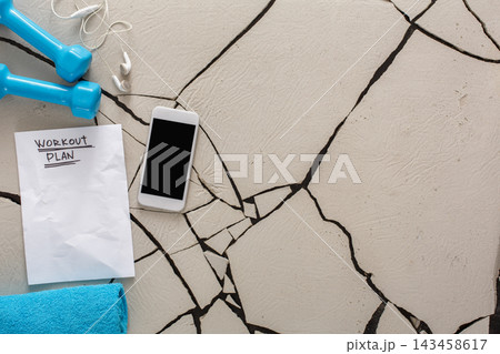 An arrangement of dumbbells, a towel, and a smartphone on a cracked surface. A piece of paper labeled workout plan lies among the items. Earphones are nearby, hinting at fitness preparation. 143458617