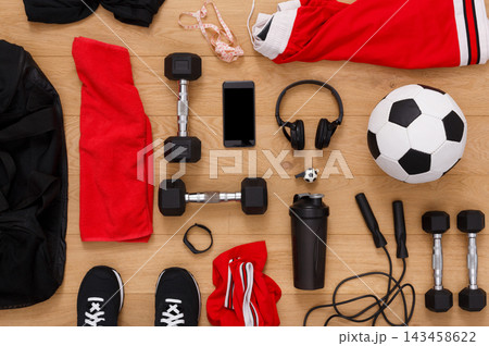Various fitness items are laid out on a wooden floor. There are dumbbells, a soccer ball, a water bottle, headphones, and workout clothes. A smartphone is also present among the gear. 143458622