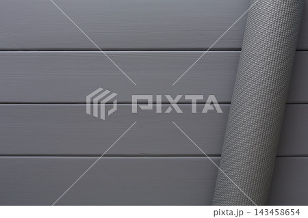 A rolled mat rests against a gray wooden floor. The floor has smooth planks arranged in neat lines. The mat has a textured surface and adds an element of design to the space. A rolled mat rests against a gray wooden floor. The floor has smooth planks arranged in neat lines. The mat has a textured surface and adds an element of design to the space. 143458654