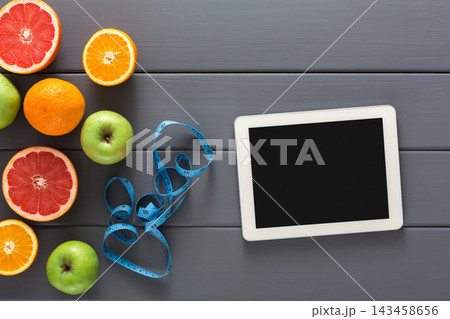 A variety of fresh fruits including oranges, apples, and grapefruits is placed on a wooden table. A measuring tape lies beside a tablet, suggesting a focus on health and diet. A variety of fresh fruits including oranges, apples, and grapefruits is placed on a wooden table. A measuring tape lies beside a tablet, suggesting a focus on health and diet. 143458656