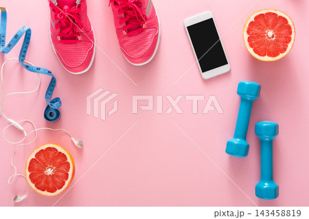 Brightly colored sports shoes are placed on a pink surface alongside dumbbells, a smartphone, and grapefruit. A measuring tape and earbuds are present, showing a fitness theme. 143458819