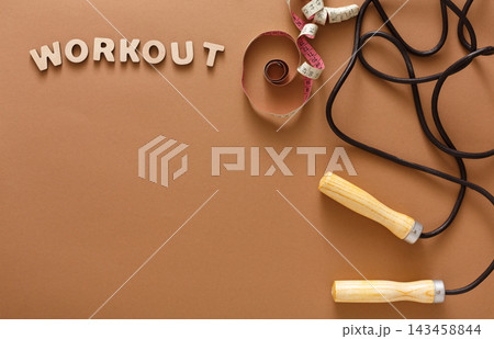 Workout items are arranged on a brown surface. There are wooden-handled jump ropes and measuring tape coiled beside letters that spell workout. The scene shows tools for fitness activities. Workout items are arranged on a brown surface. There are wooden-handled jump ropes and measuring tape coiled beside letters that spell workout. The scene shows tools for fitness activities. 143458844