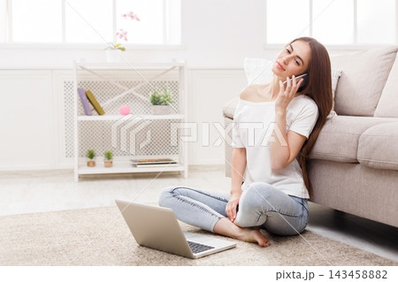 A young woman is sitting on the floor in a living room. She is talking on the phone and using a laptop in front of her. There are plants and a shelf in the background. A young woman is sitting on the floor in a living room. She is talking on the phone and using a laptop in front of her. There are plants and a shelf in the background. 143458882