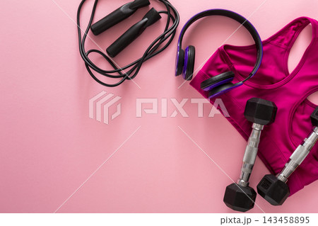 A workout outfit is placed on a pink background next to dumbbells and a jump rope. A pair of headphones is also included, suggesting preparation for a fitness session. A workout outfit is placed on a pink background next to dumbbells and a jump rope. A pair of headphones is also included, suggesting preparation for a fitness session. 143458895