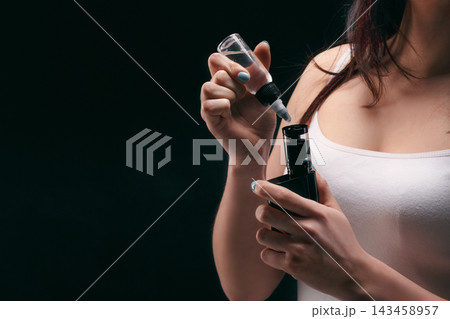 A person fills a vape device with e-liquid using a dropper. The setting is dark, and the focus is on the hands holding the vape and dropper. It is evening at home. 143458957