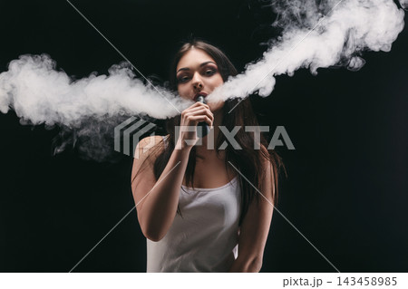 A woman stands against a dark background while exhaling vapor from a vape. She holds the device in her hand and lets out large clouds of smoke. The scene is captured in low light. 143458985
