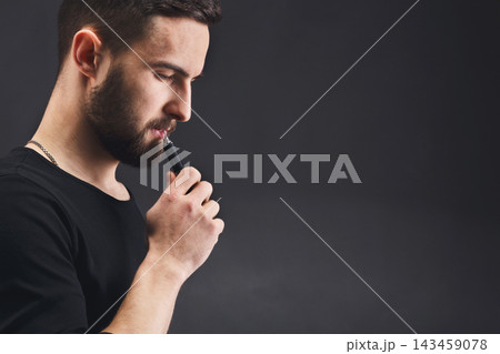 A man stands against a plain dark background, holding a vape device close to his mouth. He appears to be deep in thought. His gaze is focused, creating a moment of reflection. 143459078