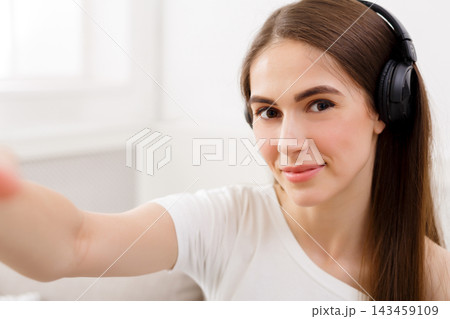 A young woman sits in a well-lit room wearing headphones. She smiles at the camera and reaches her hand out. The setting has a light, airy feel with natural light coming through the window. 143459109
