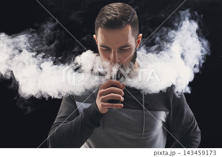 A man stands indoors, holding a vaping device. He exhales a large cloud of vapor that fills the space around him. The setting is dark, emphasizing the vapor. 143459173