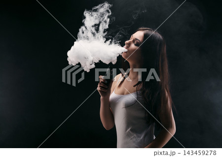 A woman exhales a thick cloud of vapor while holding a vaping device. The setting is dim with a focus on the vapor rising around her. Her long hair and casual outfit are visible. A woman exhales a thick cloud of vapor while holding a vaping device. The setting is dim with a focus on the vapor rising around her. Her long hair and casual outfit are visible. 143459278
