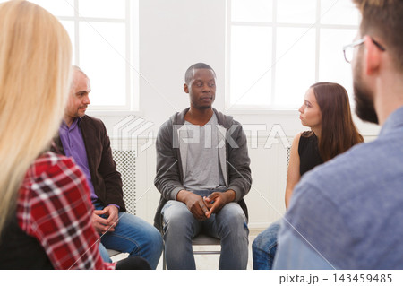 People sit in a circle in a light room. A man talks while others listen. The setting is casual and focused on sharing thoughts and experiences. 143459485