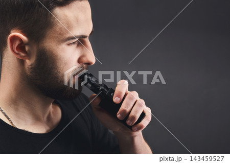 A man is using a black vaporizer indoors. He holds the device close to his mouth, focusing on the action of vaping. The background is dark, emphasizing his profile and gesture. 143459527