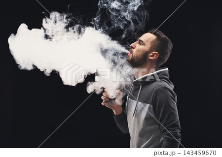 A man stands in a dark environment. He is holding a vape device and exhaling a large cloud of vapor. The scene focuses on the action of vaping and the vapor produced. A man stands in a dark environment. He is holding a vape device and exhaling a large cloud of vapor. The scene focuses on the action of vaping and the vapor produced. 143459670