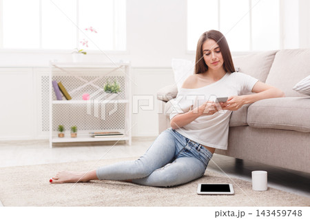 A woman sits on the floor in her living room. She is looking at her smartphone while dressed in casual clothes. The space has a couch, a shelf, and a small table nearby. A woman sits on the floor in her living room. She is looking at her smartphone while dressed in casual clothes. The space has a couch, a shelf, and a small table nearby. 143459748