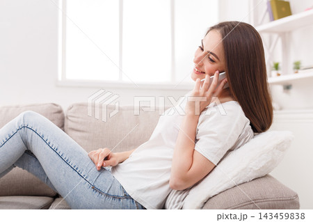 A young woman is sitting on a couch in her home, talking on her phone with a smile. Natural light enters through a window. She sits comfortably and seems engaged in her conversation. A young woman is sitting on a couch in her home, talking on her phone with a smile. Natural light enters through a window. She sits comfortably and seems engaged in her conversation. 143459838