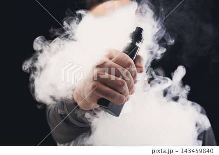A person uses a vape device and exhales a large cloud of vapor. The setting is indoors with dim lighting. The focus is on the device and vapor, creating a secluded atmosphere. 143459840