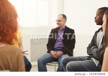 Five people are seated in a circle in a well-lit room. They engage in a discussion, with one person speaking and others listening attentively. 143459948