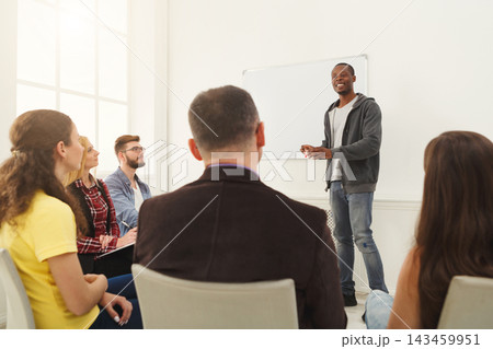 A speaker stands in front of a group during a workshop. Participants sit in chairs, listening and engaging. The room has large windows and a whiteboard. 143459951