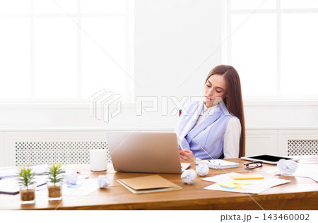 A woman sits at a desk in an office space. She looks focused as she writes notes while surrounded by crumpled papers. A laptop and notebooks are on the desk under bright light. 143460002