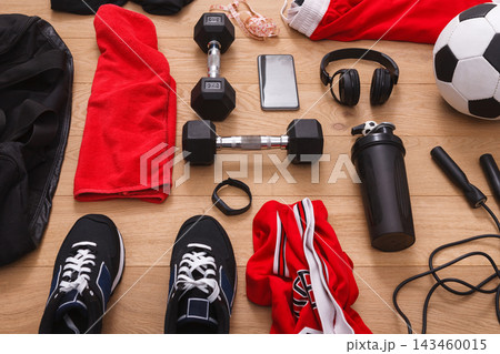 Workout gear and equipment is arranged on a wooden floor. Items include dumbbells, a water bottle, sneakers, a phone, headphones, and clothing items for exercise. 143460015