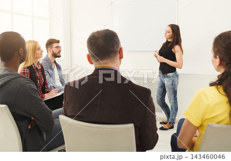 A woman stands in front of a group of people in a classroom. The group appears engaged as they listen to her speak. The setting has bright natural light coming from windows. 143460046