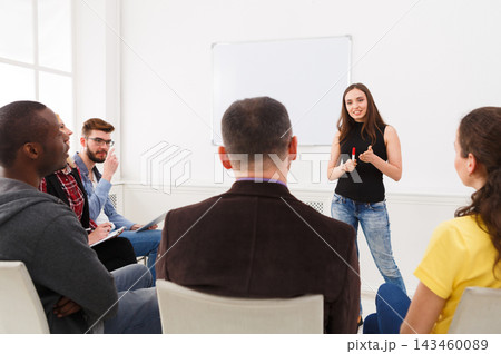 A speaker stands in front of a small group in a bright room. The audience looks engaged and attentive while the speaker uses gestures to explain points. Notebooks and pens are visible. 143460089