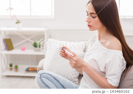 A woman is sitting on a chair in a well-lit room. She looks at a thermometer in her hand. Green plants can be seen in the background along with some books and decor. A woman is sitting on a chair in a well-lit room. She looks at a thermometer in her hand. Green plants can be seen in the background along with some books and decor. 143460102
