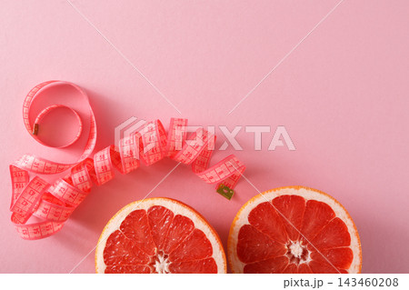 Two halves of a grapefruit sit on a pink surface, with a measuring tape curled beside them. This scene relates to health and nutrition themes, focusing on diet and wellness. 143460208