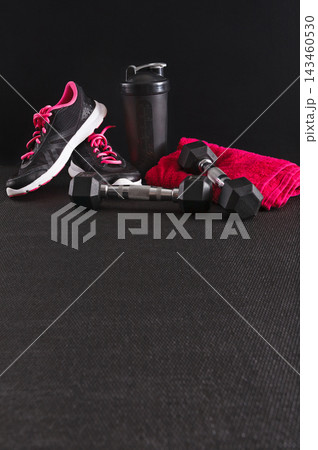 A pair of black and pink sports shoes rests beside dumbbells and a red towel. A shaker bottle is nearby. This setup suggests preparation for a workout at a gym or home. A pair of black and pink sports shoes rests beside dumbbells and a red towel. A shaker bottle is nearby. This setup suggests preparation for a workout at a gym or home. 143460530