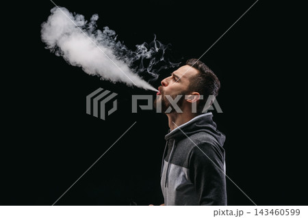 Man stands in a dark space and exhales a large cloud of vapor. Smoke rises into the air, creating a dramatic effect. His expression shows focus on the action he is performing. Man stands in a dark space and exhales a large cloud of vapor. Smoke rises into the air, creating a dramatic effect. His expression shows focus on the action he is performing. 143460599