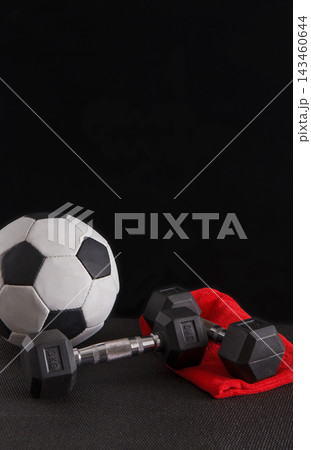 A soccer ball sits next to two dumbbells on a dark surface. A red cloth lies in the scene, adding a splash of color. The setup suggests a focus on fitness and sports activities. 143460644