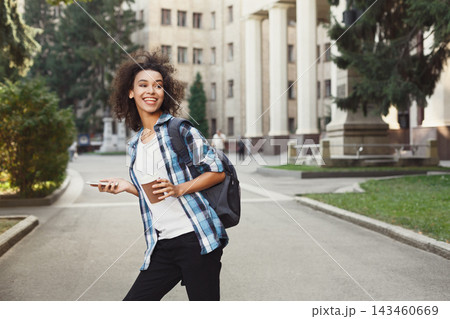 A young woman walks on a sunny day carrying a coffee cup and a phone. She looks happy while strolling through a college campus with buildings and trees around her. 143460669