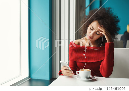 A woman is seated in a cafe, looking at her smartphone with a coffee cup in front of her. She has curly hair and appears to be in thought or feeling stressed. Natural light comes through the window. A woman is seated in a cafe, looking at her smartphone with a coffee cup in front of her. She has curly hair and appears to be in thought or feeling stressed. Natural light comes through the window. 143461003