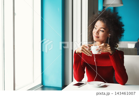A woman sits in a cafe holding a cup of coffee while enjoying music through her earphones. She looks thoughtful as she gazes out the window. The cafe features a bright blue wall. 143461007