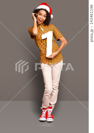 A young person smiles while wearing a Santa hat and holding up the number one. The scene is colorful and festive, capturing a moment of joy. The backdrop is a simple gray. A young person smiles while wearing a Santa hat and holding up the number one. The scene is colorful and festive, capturing a moment of joy. The backdrop is a simple gray. 143461196