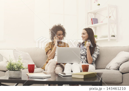 Two friends sit on a couch in a bright living room. One friend talks on the phone while the other uses a laptop. There are mugs on the table. They share smiles and moments. 143461210