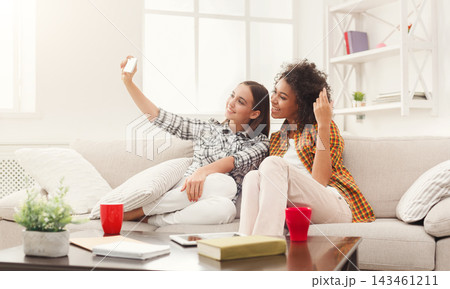 Two friends are sitting on a couch in a cozy room. They are smiling and taking a selfie together. Bright light enters from the window. They are enjoying a fun moment together. 143461211