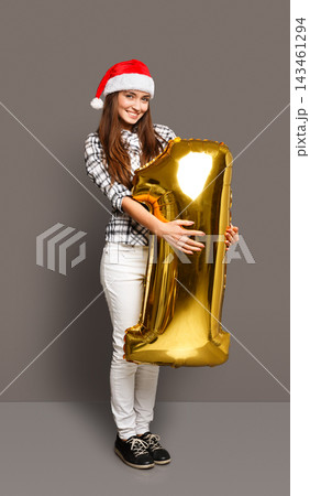 A woman stands against a gray background wearing a santa hat. She holds a large gold number one balloon while smiling. This scene shows a festive moment, likely for a birthday or new year. A woman stands against a gray background wearing a santa hat. She holds a large gold number one balloon while smiling. This scene shows a festive moment, likely for a birthday or new year. 143461294