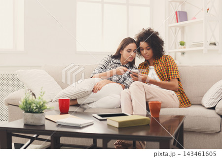 Two friends sit closely on a couch in a well-lit room. They look at a mobile device while sharing a moment. Nearby, there are cups and books on a table. The setting feels relaxed and casual. 143461605
